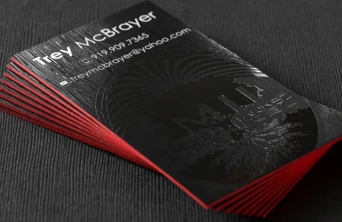 Stacked black silk business cards for Trey McBrayer rest on a dark textured surface, featuring glossy spot UV graphics and vibrant red painted edges.