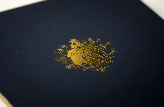 A square dark blue silk business card rests on white, featuring a gleaming gold foil botanical emblem in the center.