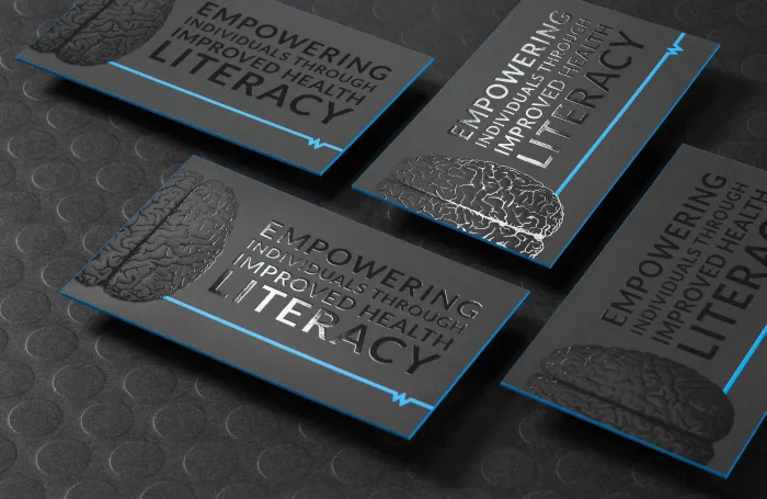 Four black silk health literacy business cards rest on a dark textured surface, featuring raised silver foil brain graphics, lettering.