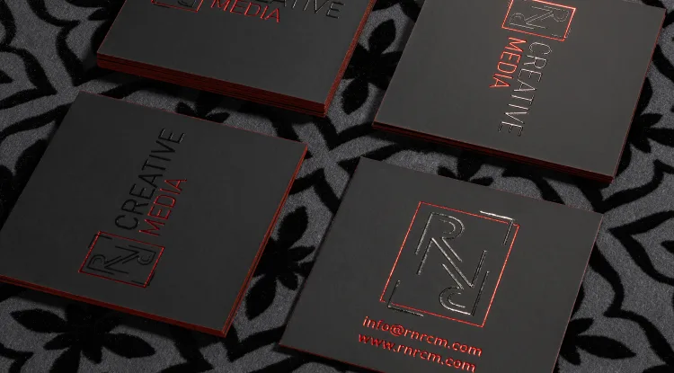 Square black suede business cards for RNRC Creative Media rest on patterned gray fabric, featuring gleaming red foil lettering and metallic red edges.