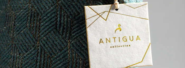 A white hang tag with gold foil geometric line-art and Antigua Collection lettering, hanging by a cord from a teal woven-texture fabric.