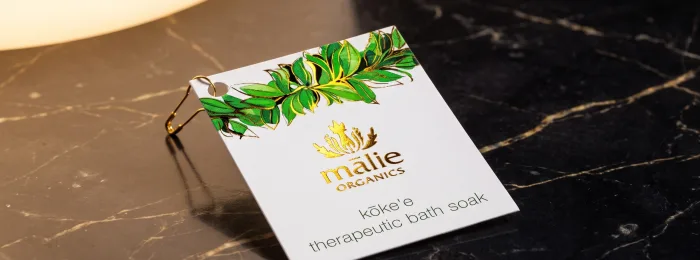A white, gold-foil hang tag for Mālie Organics with a green and gold foil laurel-leaf branch across the top, clipped on with a small gold pin.