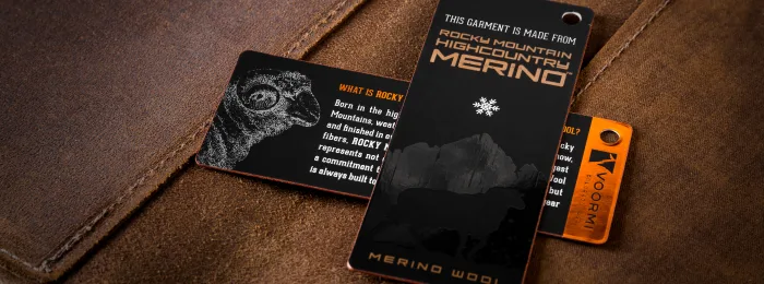 Three foil hang tags on a leather-like brown surface: a black tag with a ram graphic, a black tag reading 'Rocky Mountain Highcountry Merino,' and an orange tag with a snowflake logo.