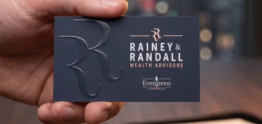 A hand holds a Rainey and Randall wealth advisors navy business card featuring a large debossed RR monogram and rose gold foil lettering.