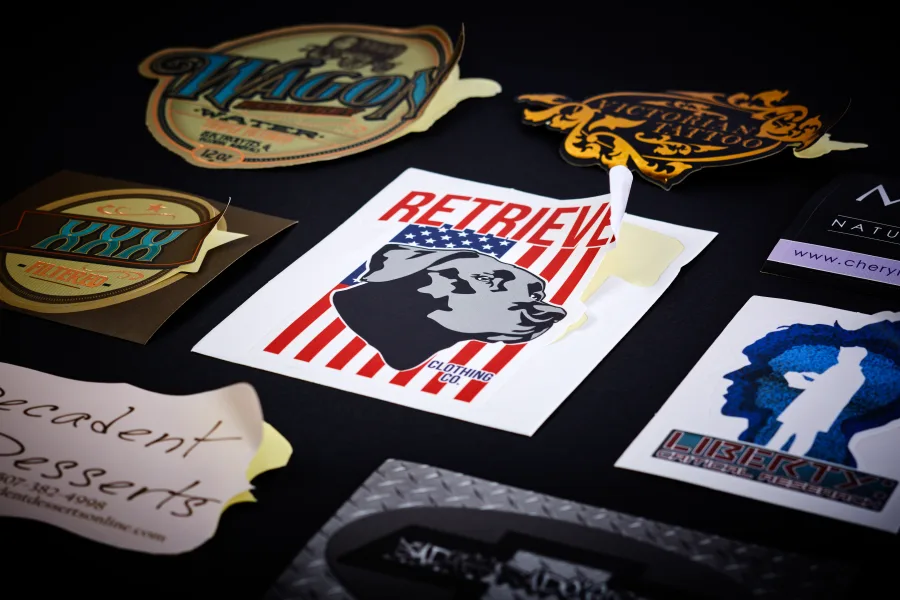 A flat lay of custom die-cut vinyl label stickers, each cut to the exact silhouette of its own badge artwork, on a leather surface.