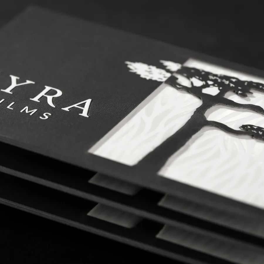 A black and white multilayer business card for Ayra Films features a die-cut tree silhouette revealing a textured white interior layer with raised lettering.